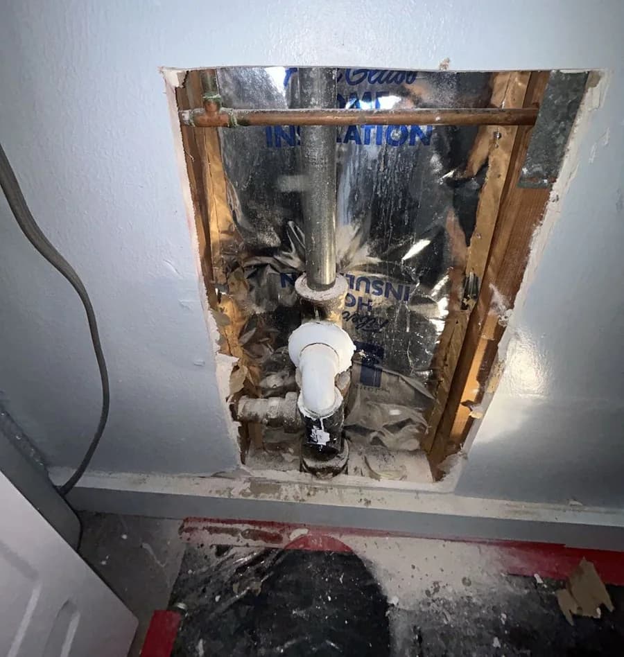 An opened wall cavity showing a copper supply line and a galvanized stack running down to a plastic trap fitting